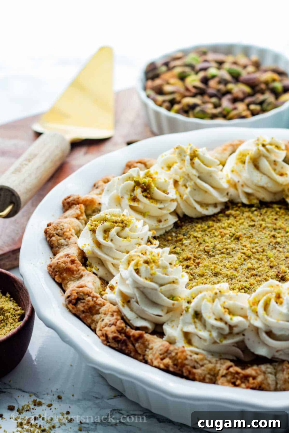 Velvet Pistachio Dream Pie 2 A beautifully baked Pistachio Custard Pie in a white pie pan, with a pie server and a small bowl of fresh pistachios beside it, set on a rustic wooden surface.
