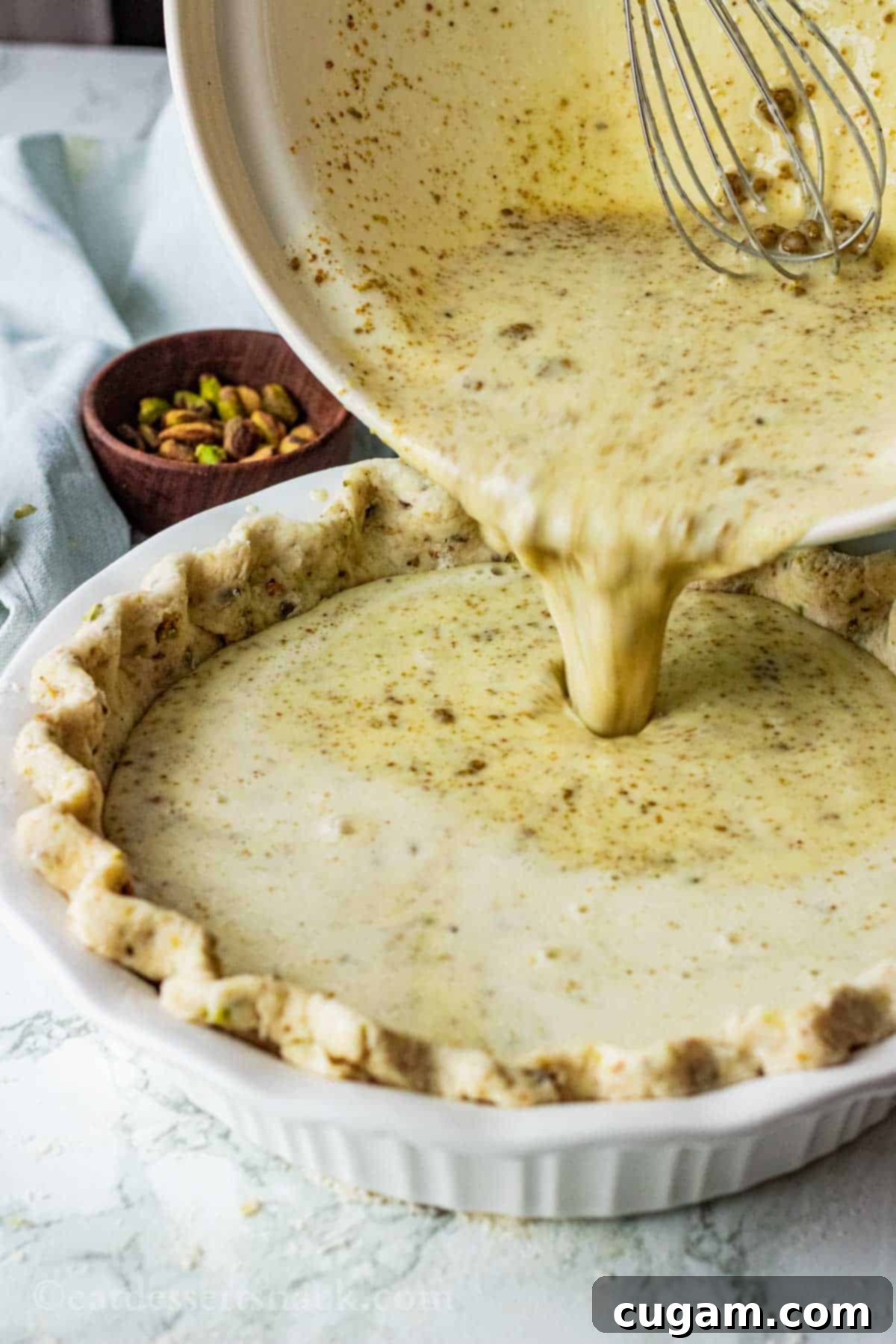 Velvet Pistachio Dream Pie 12 A bowl of creamy pistachio custard being carefully poured into a raw, crimped pistachio pie crust set in a white pie dish.