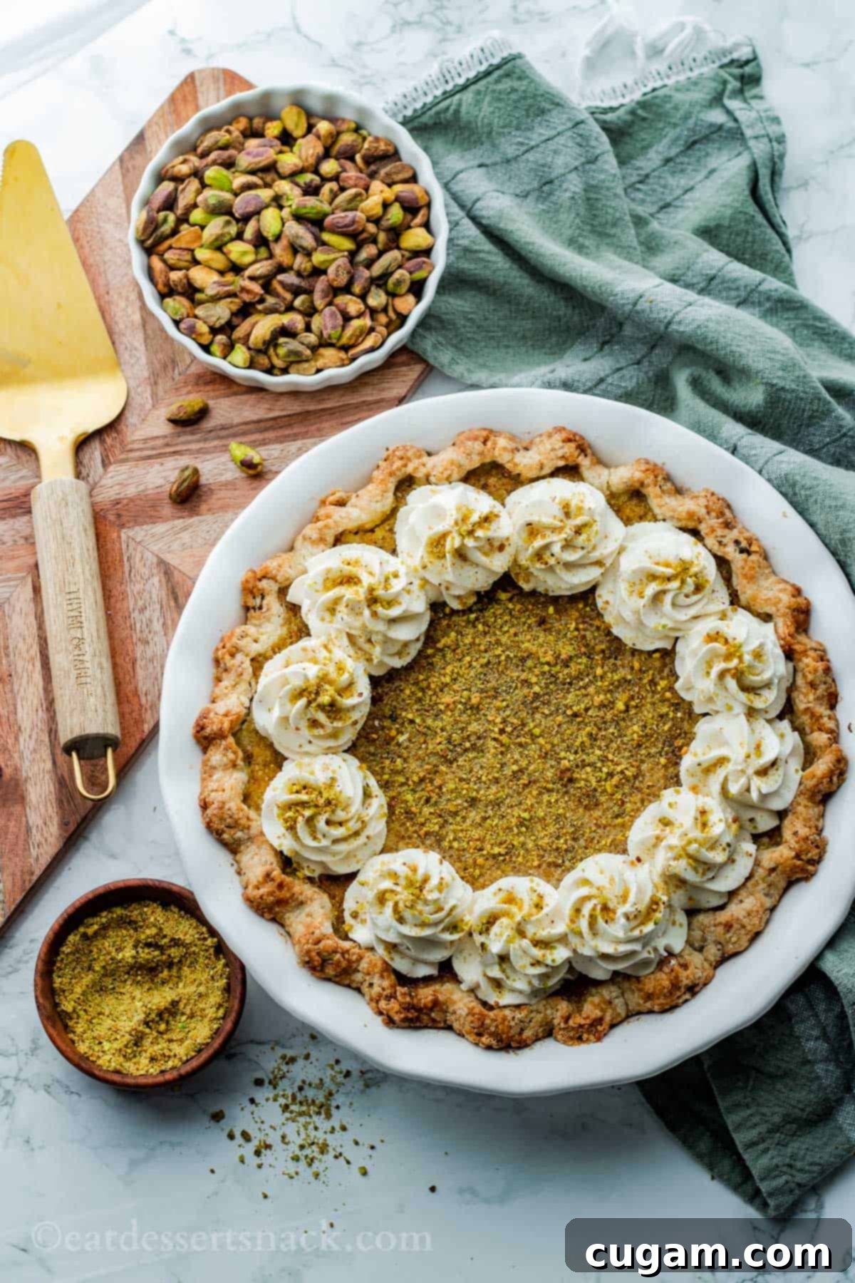 Velvet Pistachio Dream Pie 13 A fully baked pistachio pie in a white pie dish, with a pie server and a bowl of pistachios on a green dish towel, all on a marble countertop.