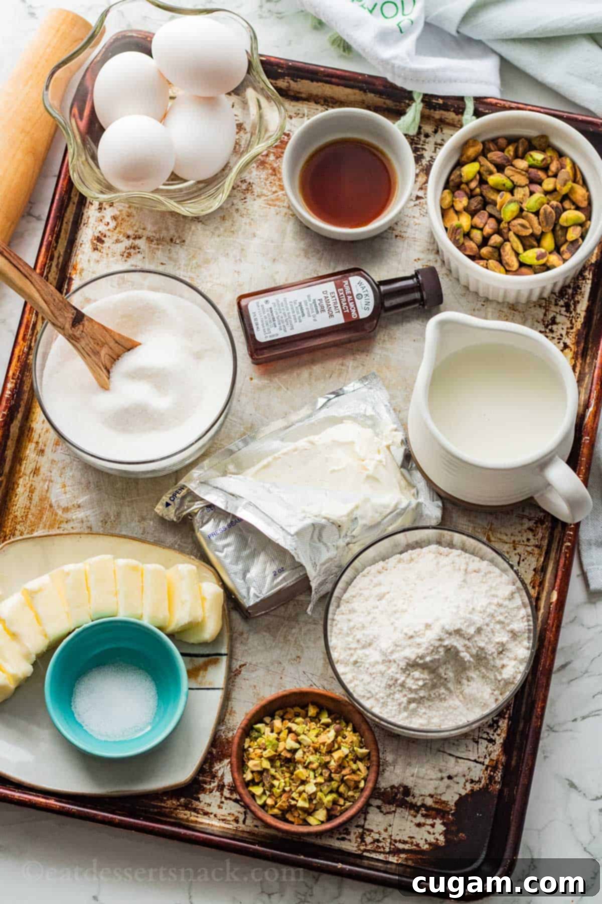 Velvet Pistachio Dream Pie 3 An array of fresh ingredients for Pistachio Custard Pie laid out on a metal baking pan, including flour, butter, salt, pistachios, cream cheese, eggs, vanilla, almond extract, and milk in various bowls.