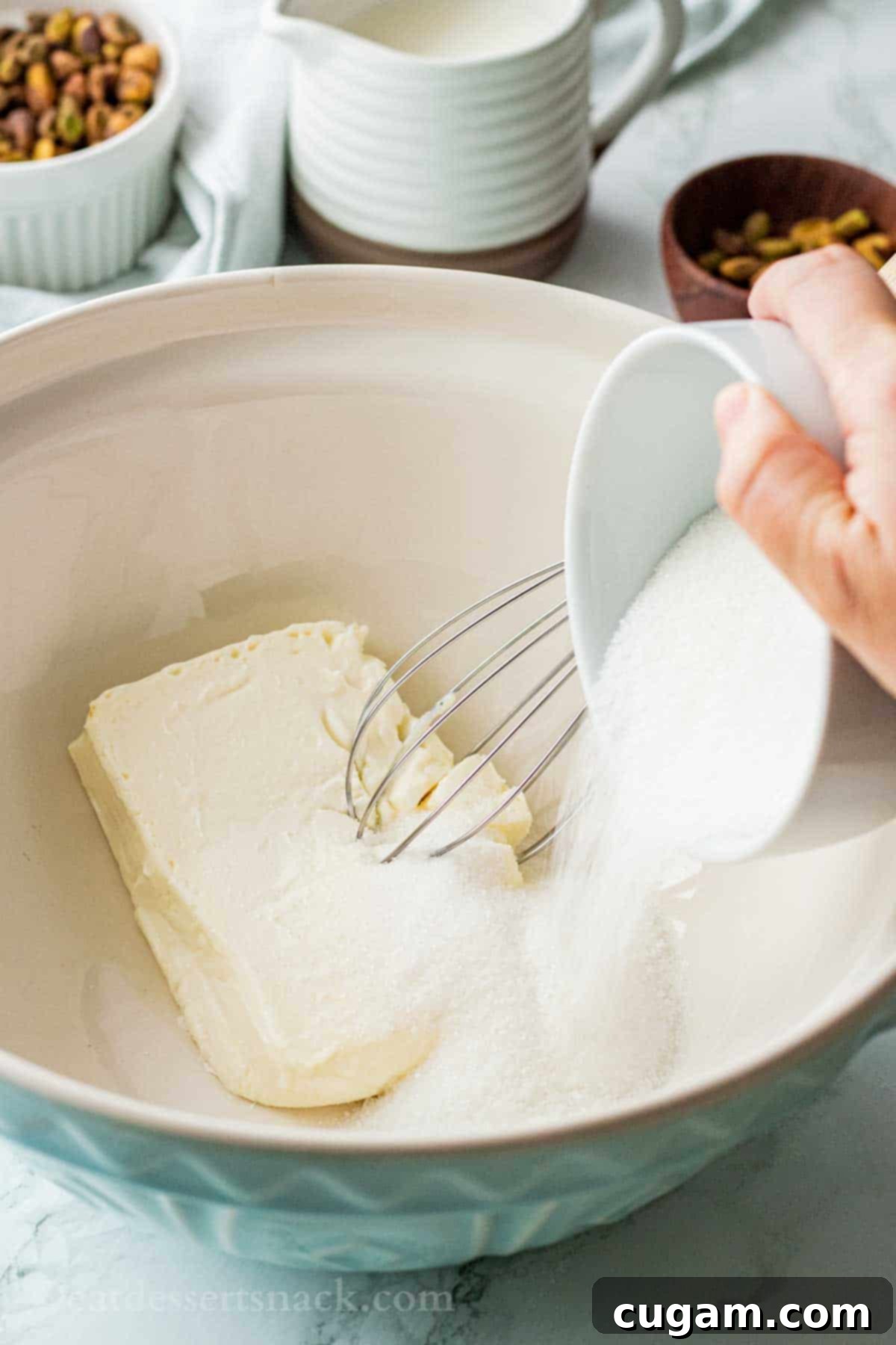 Velvet Pistachio Dream Pie 8 A hand pouring white granulated sugar from a white bowl into a blue mixing bowl containing softened cream cheese, with a whisk nearby.