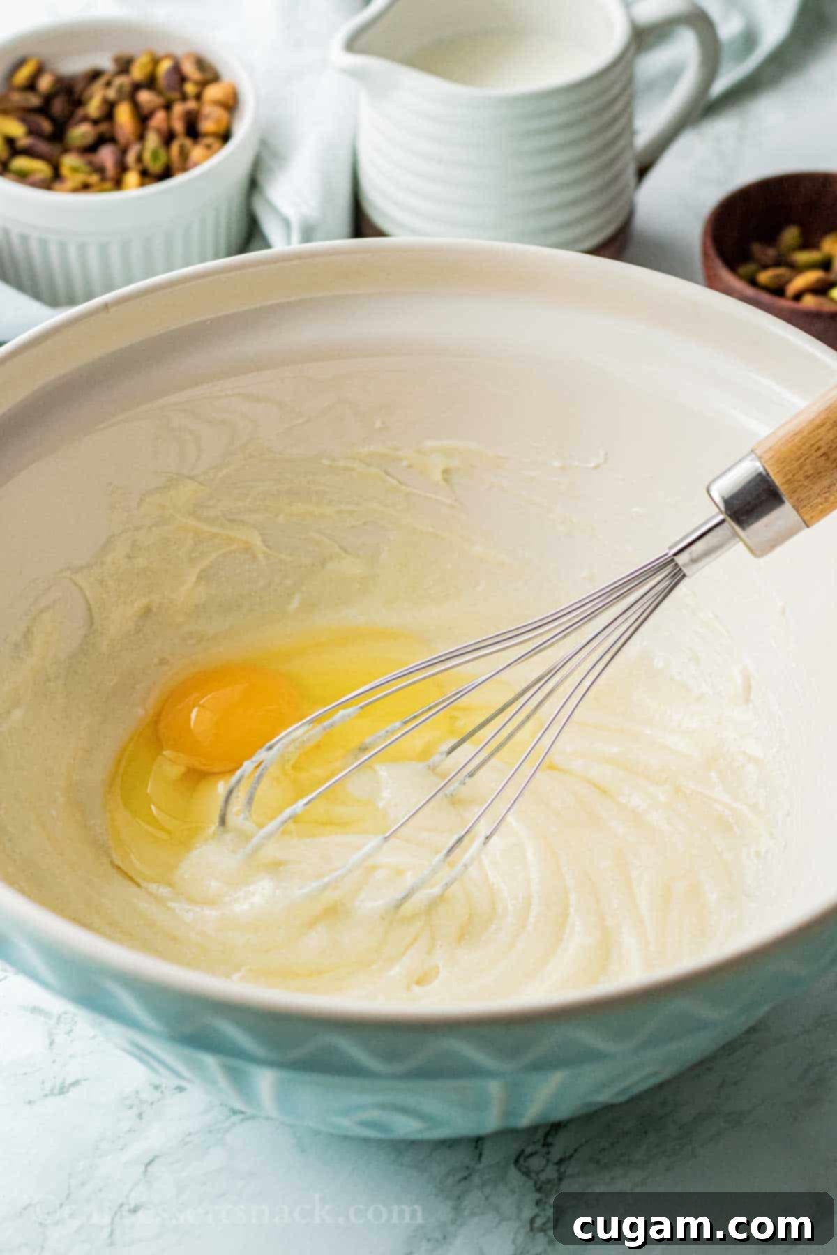 Velvet Pistachio Dream Pie 9 A whisk in a blue bowl, mixing an egg into a creamy mixture of cream cheese and sugar.