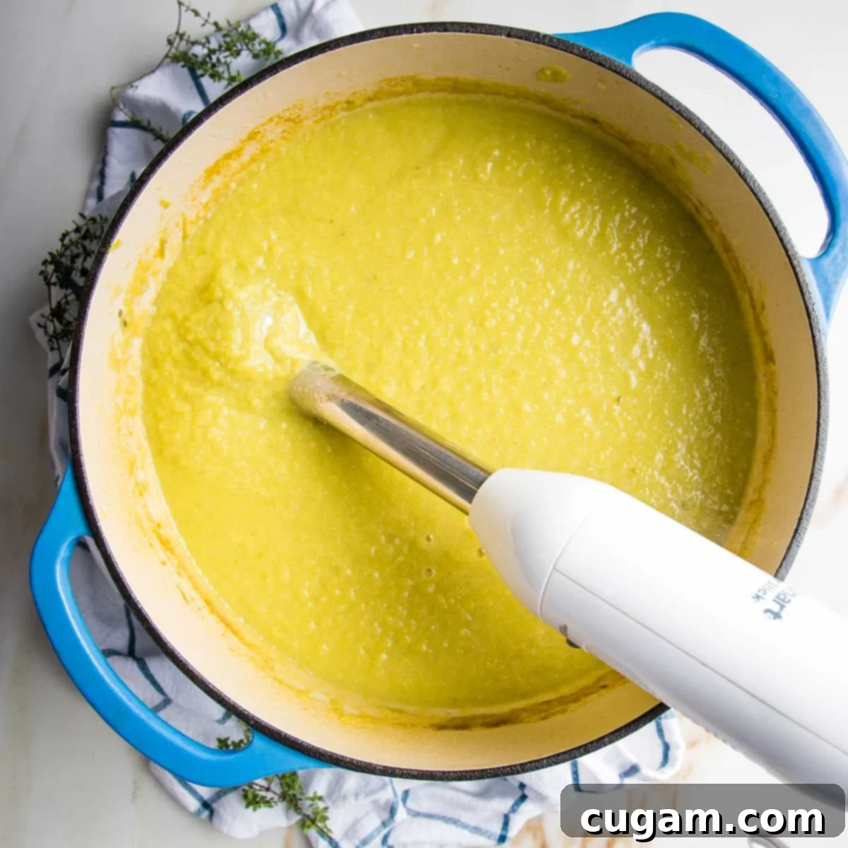 An immersion blender is shown in a Dutch oven, actively pureeing the cooked vegetable soup into a smooth, creamy consistency. Steam rises from the pot, indicating the soup is still hot.