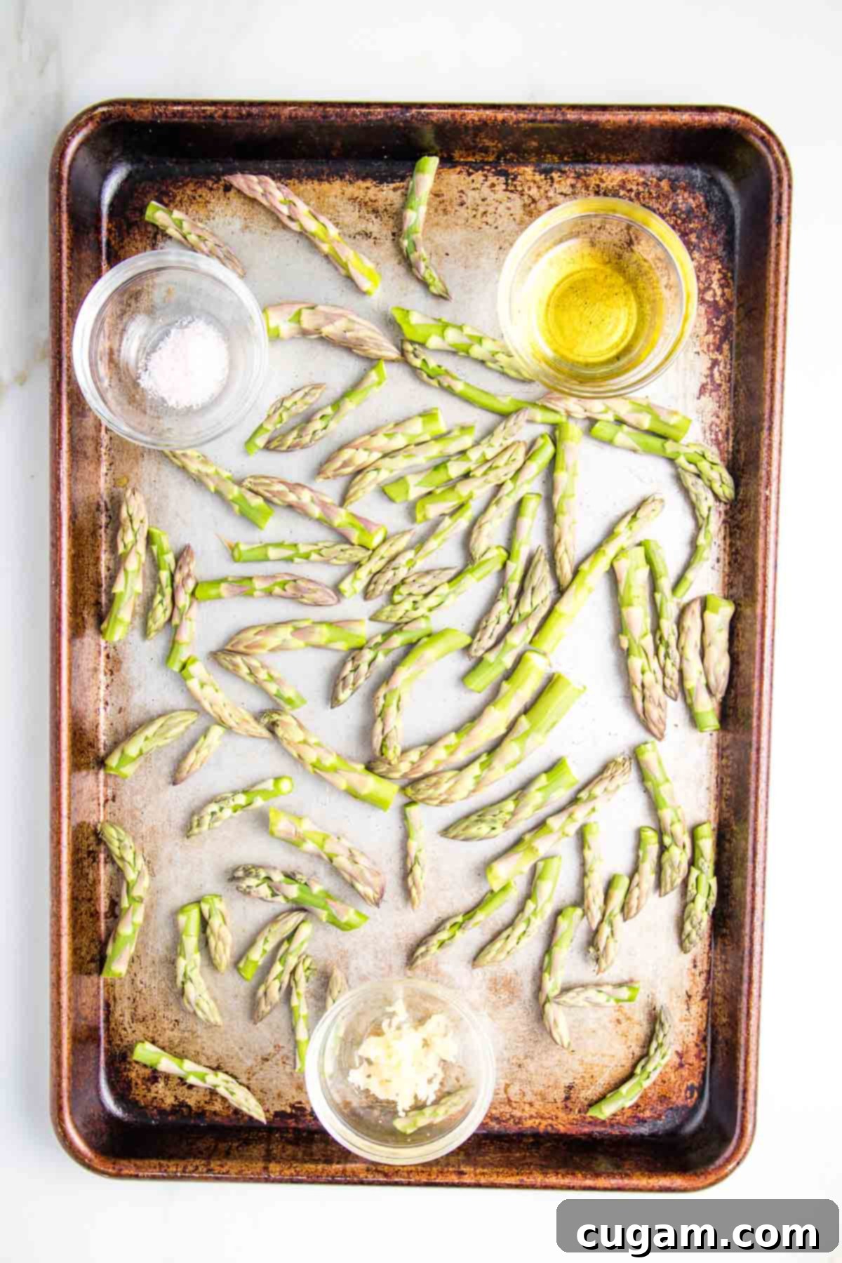 A baking tray holds raw asparagus tips, surrounded by small dishes containing seasoning, minced garlic, and olive oil, all prepared for roasting.