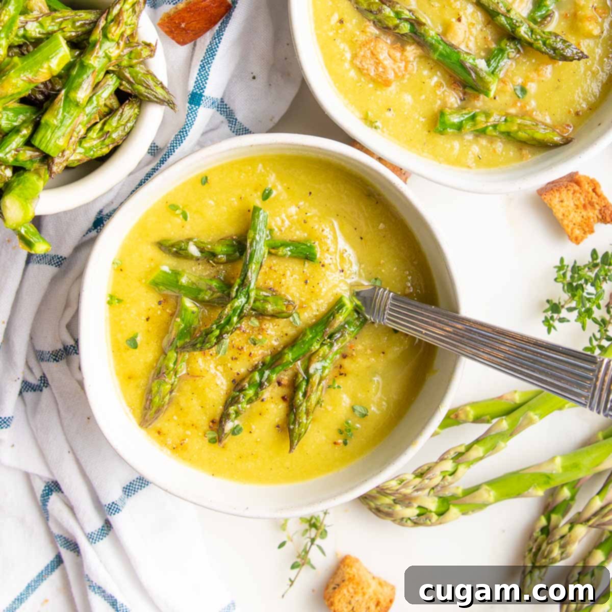 Several bowls of creamy yellow vegan asparagus soup are garnished with garlicky asparagus tips. The counter around the bowls features a white and blue dish towel sprinkled with croutons, a small dish of roasted asparagus, and some fresh, raw asparagus spears, creating an inviting culinary scene.