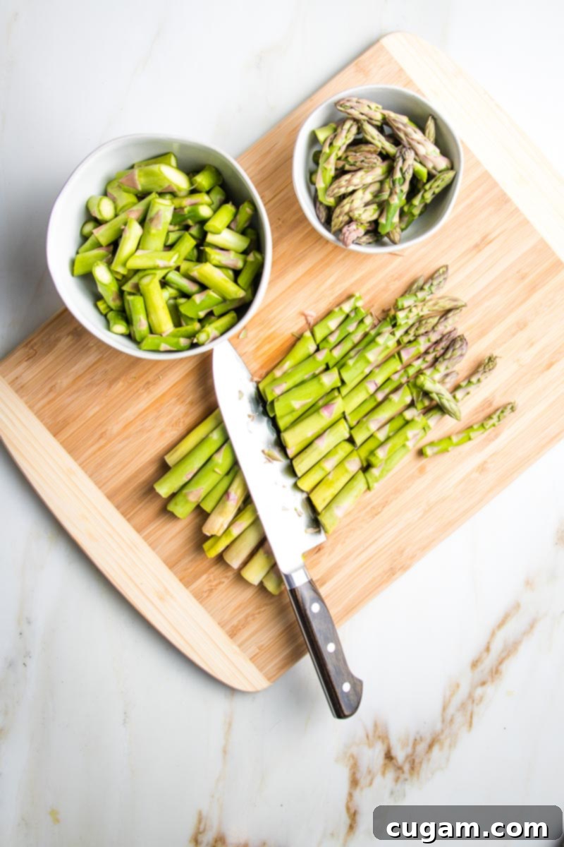 A bamboo cutting board holds a bunch of asparagus, with a chef's knife. The asparagus is cut into 1-inch pieces, separated into two bowls: a smaller one for the tips and a larger white bowl for the remaining stalk pieces.