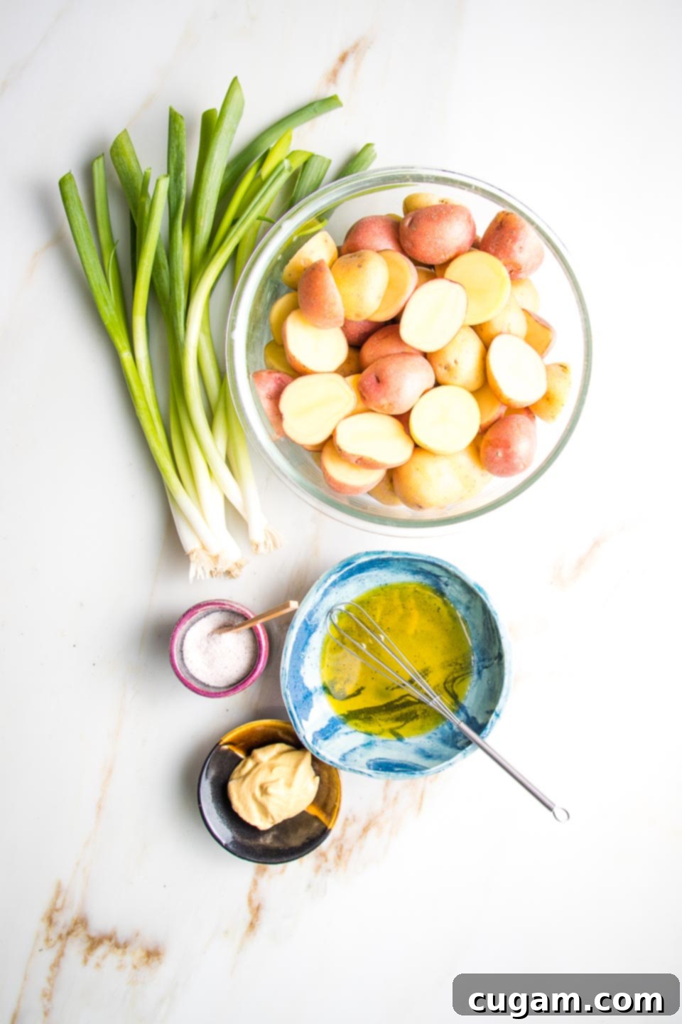 The five essential ingredients for grilled potato salad: fresh scallions, small potatoes, vibrant mustard, olive oil, and coarse sea salt arranged on a rustic wooden surface.