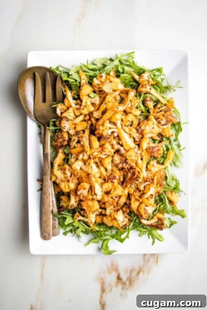 Large white rectangular platter, scattered with fresh arugula and then generously topped with paprika roasted cauliflower, with bronze serving pieces on the left side of the plate, ready to be served.