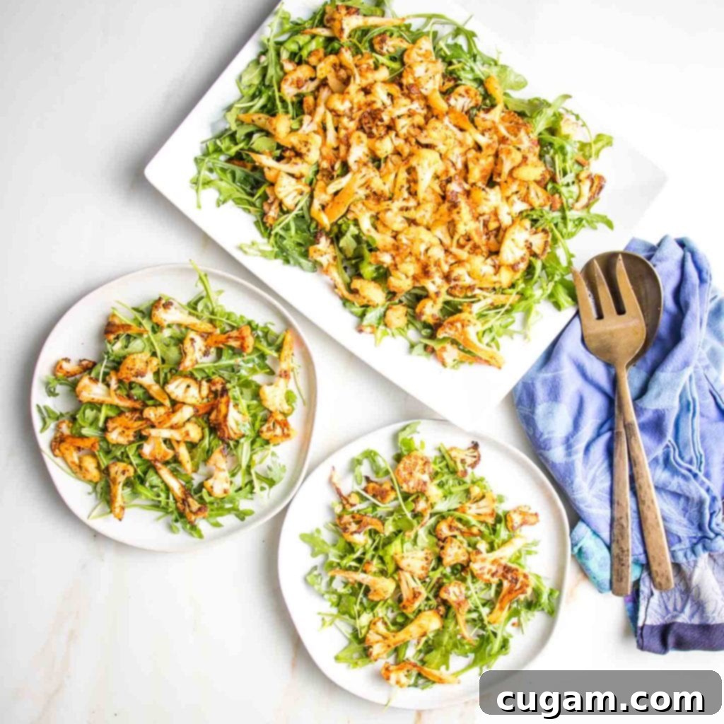 Large rectangular plate filled with vibrant arugula and generously topped with roasted cauliflower. Two additional circular rimmed plates hold individual servings of the same delicious cauliflower salad, with bronze serving pieces resting off to the side on a blue tea towel, highlighting a healthy and appealing meal.
