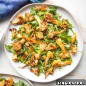 Close-up of a white plate, showcasing vibrant green arugula and perfectly roasted golden-brown cauliflower with paprika seasoning. The edge of a blue napkin is visible in the upper left corner, adding a touch of color.