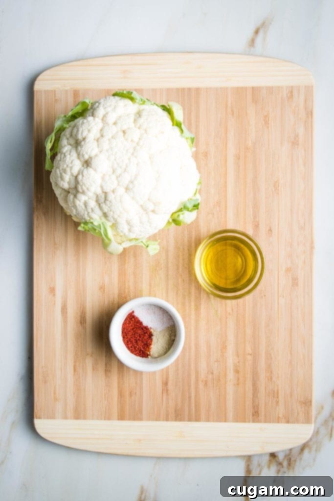 Wooden cutting board displaying a fresh whole head of cauliflower, a small glass dish containing golden olive oil, and a small white dish holding a mixture of paprika, coarse sea salt, and white pepper, all prepared for roasting.
