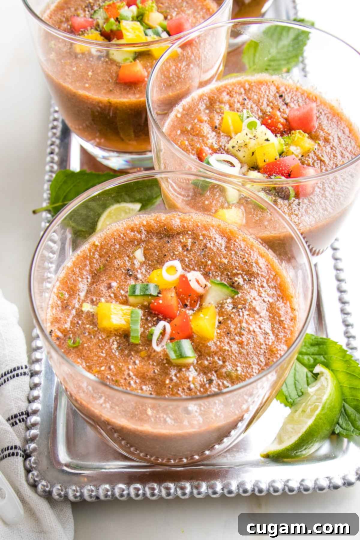 silver tray with glasses of watermelon gazpacho with chunks of colorful veggies floating on top. garnishes of fresh mint and lime wedges on the tray.
