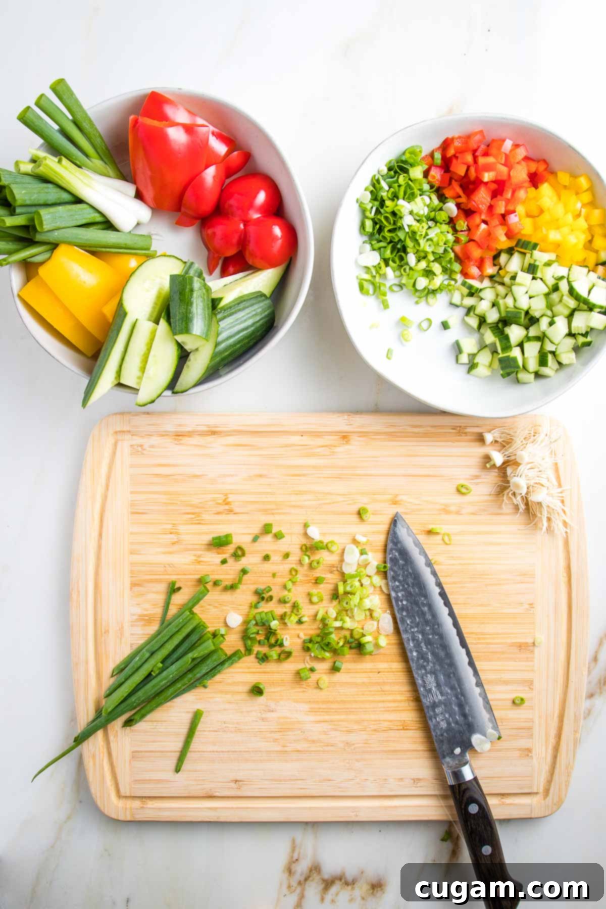 Thinly slicing scallions on a wooden cutting board. Above are 2 white bowls, one with chunks of bell peppers, scallions and cucumbers, the other has the same veggies in small dice.