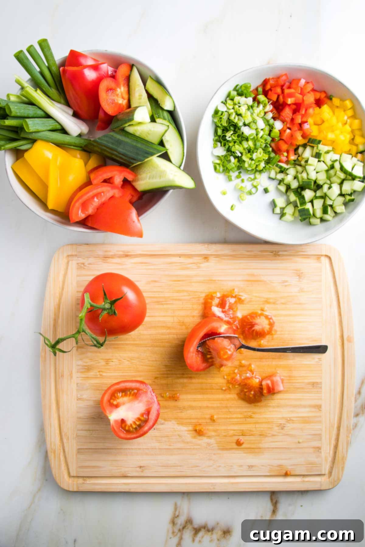 Cutting board shows a tomato on the vine and another that has been cut in half and the seeds are being scooped out with a grapefruit spoon.  There are two white bowls above the cutting board, one with chunks of colorful veggies and the other with small diced yellow and red peppers, cucumbers and scallions