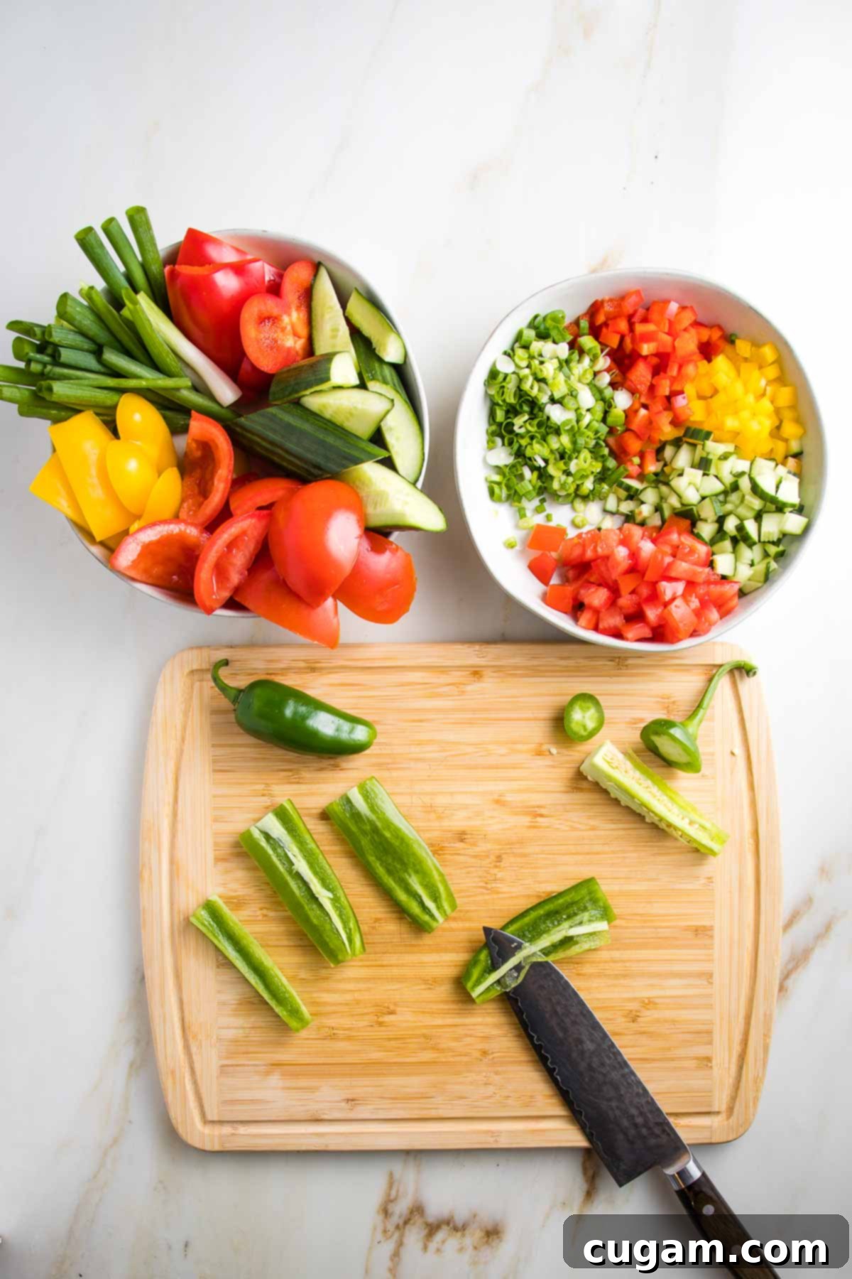 Two bowls, one filled with chunky vegetables, the other the veggies are diced small: red and yellow peppers, cucumbers, scallions, tomatoes. On the cutting board a jalapeno is trimmed and the membranes are being removed from slices.