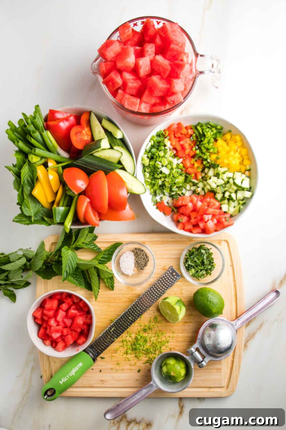 Everything ready to make watermelon gazpacho. A bowl of cubed watermelon, a bowl of chunky veggies to puree for the soup, a bowl with small dice veggies to add after, some fresh mint leaves a zested lime and a juicer, plus salt and pepper. 