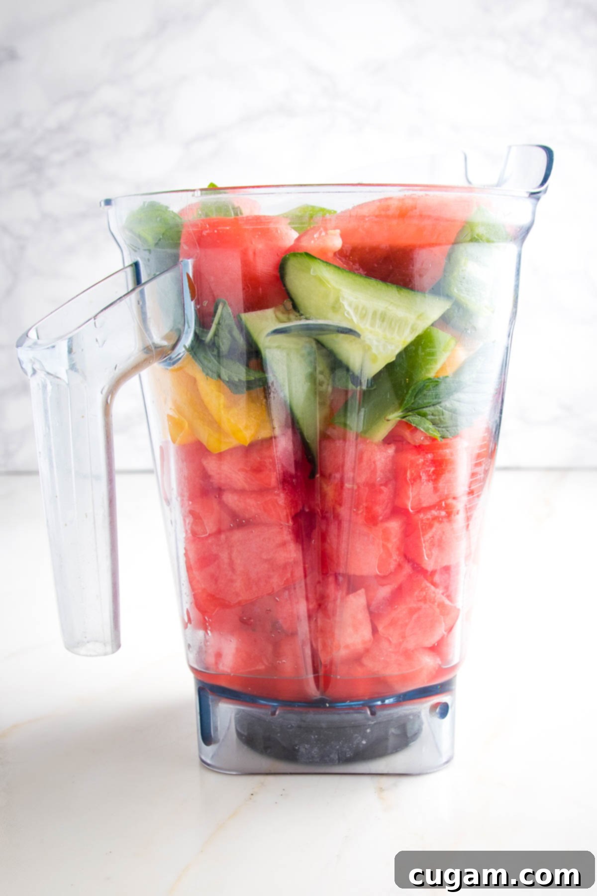 Close up of a pitcher filled with chunks of watermelong, red and yellow peppers, cucumbers, tomatoes, scallions and fresh mint leaves. In a vitamix blender pitcher about to be blended into gazpacho.