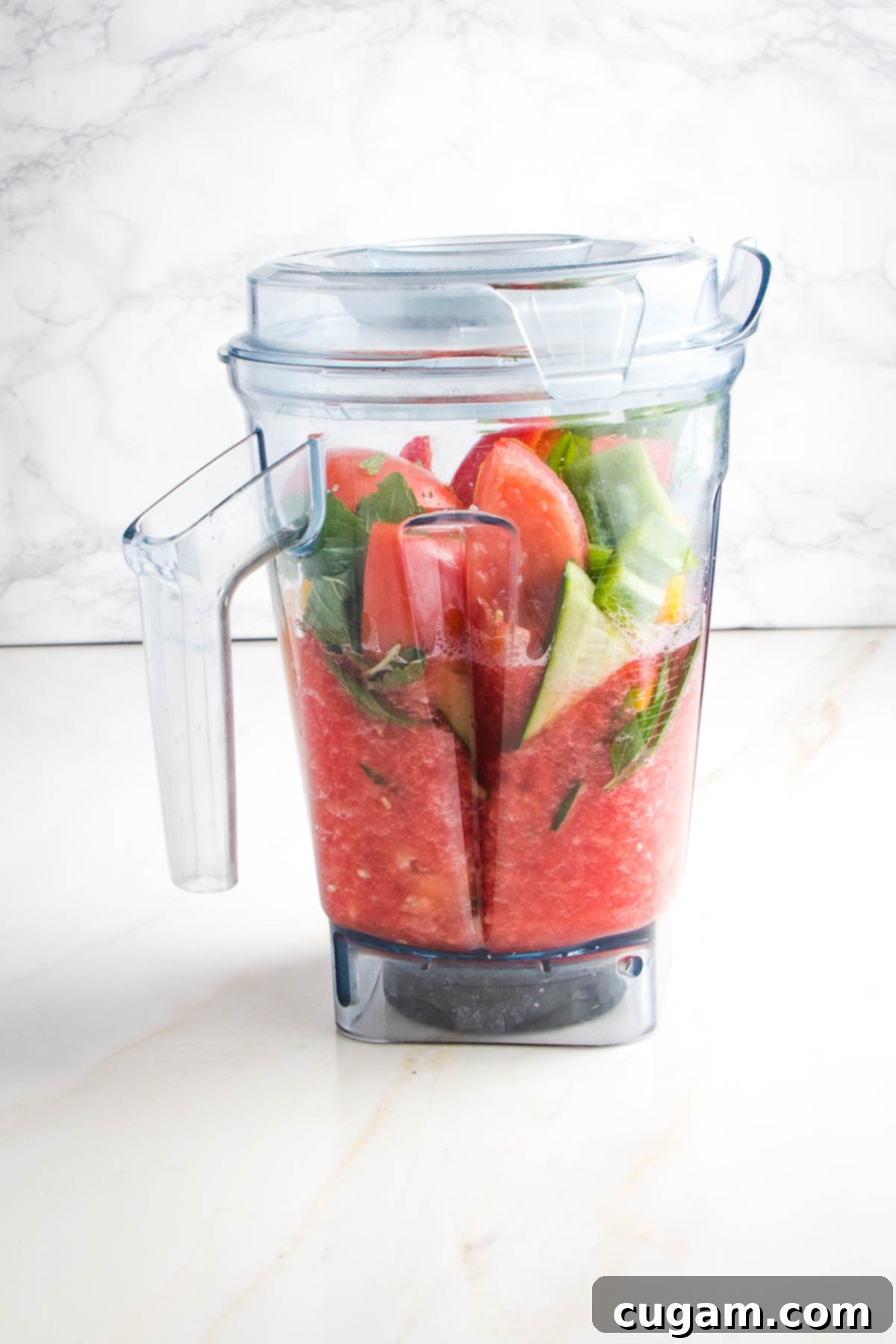 Vitamix pitcher with half blended watermelon gazpacho. The bottom is blended in a pinkish red color and on the top are ingredients yet to be blended...chunks of tomato, cucumbers, watermelon and mint leaves.