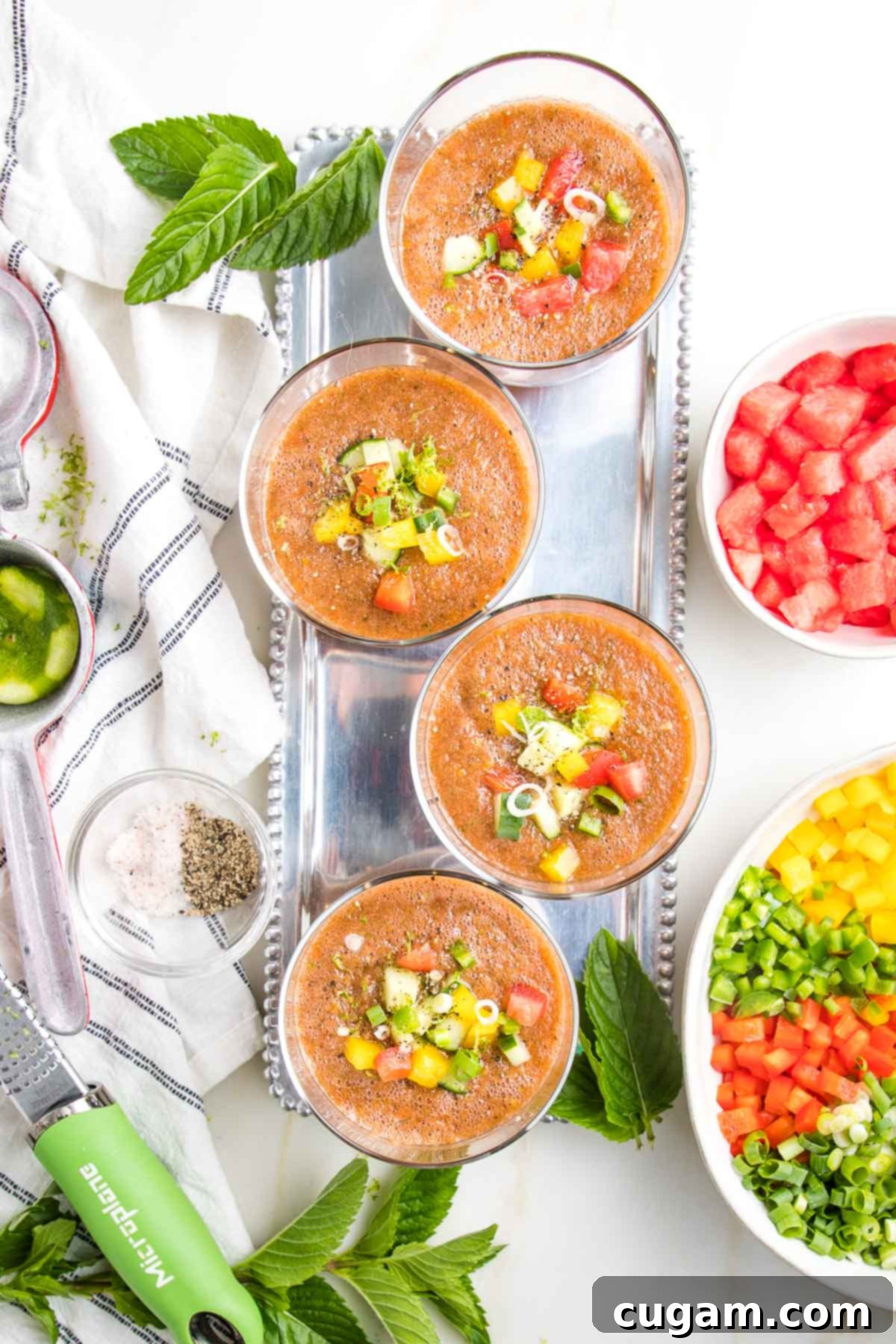 Watermelon Gazpacho in 4 glasses with chunks of colorful veggies floating on top. There is a bowl of cut up veggies on the side, and a bowl of watermelon chunks.  On the other side is a white towel with blue stripes, some lime zest and wedges, and fresh mint leaves.
