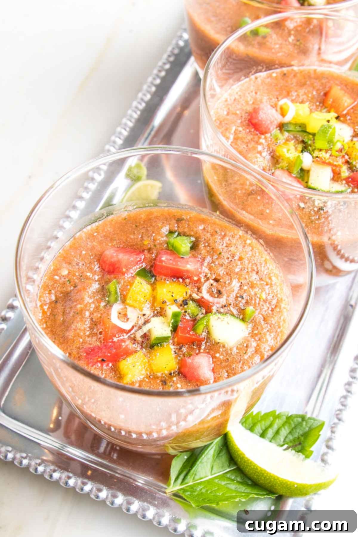 Close up of a glass of watermelon gazpacho filled with chunks of diced colorful veggies...on a silver tray with more glasses of gazpacho and some fresh mint and lime wedges.