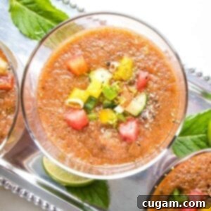 Close up of a glass filled with watermelon gazpacho with small diced, colorful veggies and cracked black pepper. Glass is on a silver tray with other glasses of gazpacho and also some mint leaves and slices of lime.