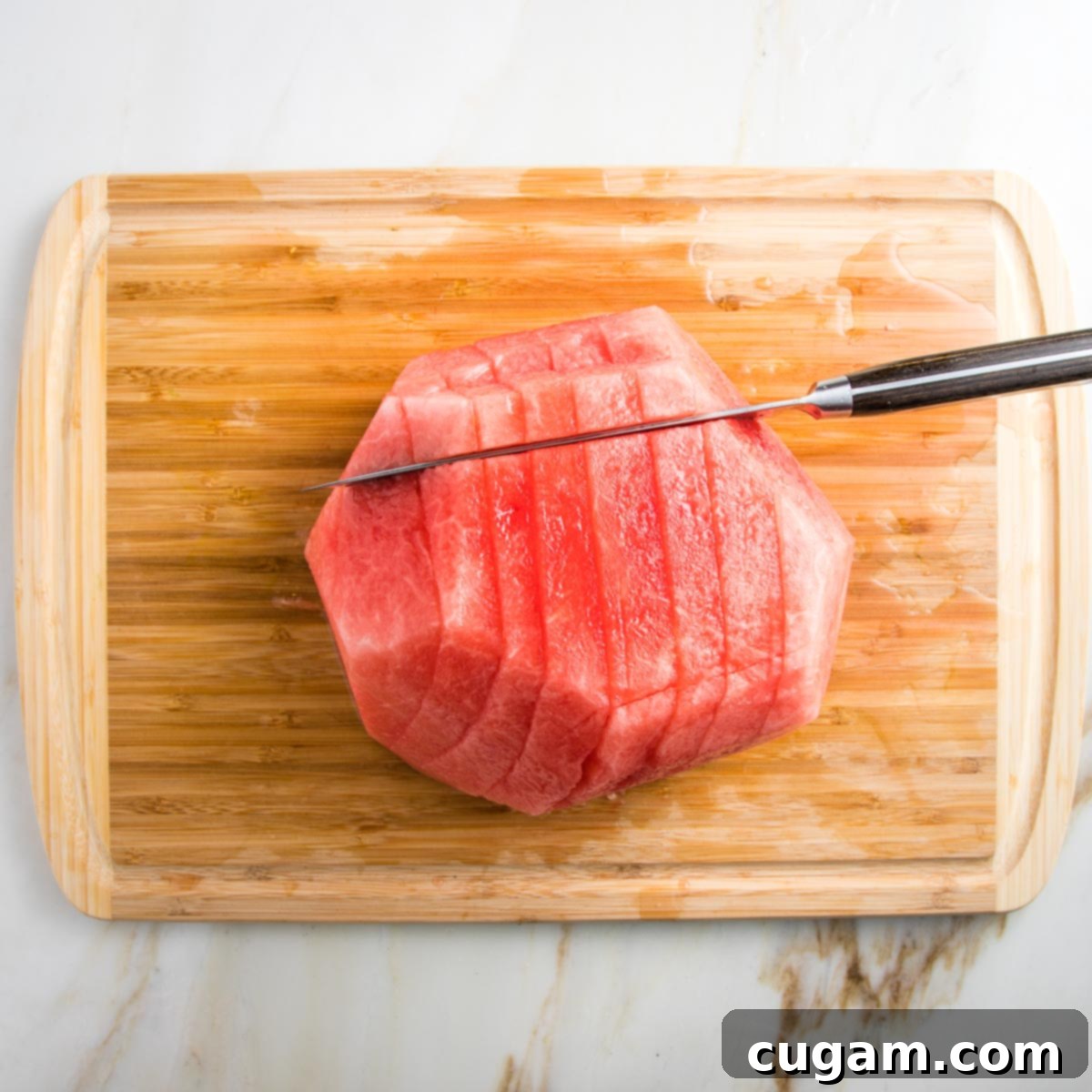 wooden cutting board. half a watermelon cut side down, aready has rind removed. cut into long slices and now slicing the other direction.