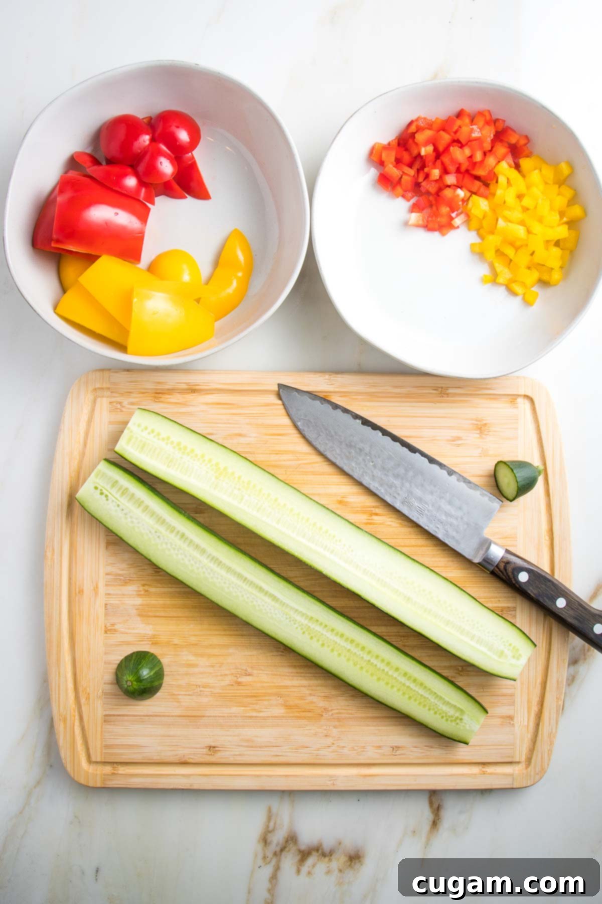 two white bowls, one with rough chop bell peppers, one with minced yellow and red peppers. Also a bamboo board with a halved enblish cucumber and chef knife.