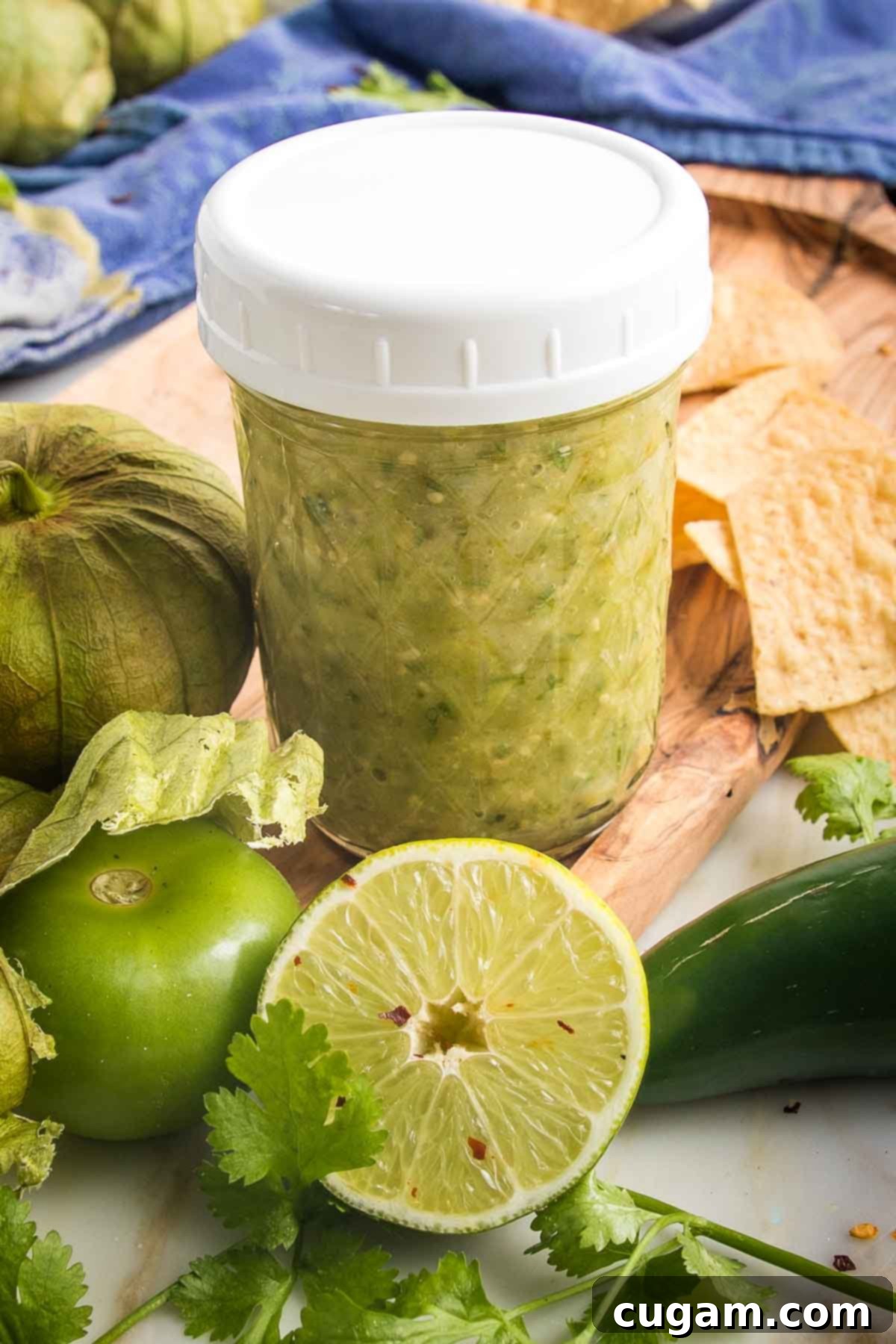 Zesty Roasted Tomatillo Salsa 13 Small mason jar filled with green salsa, surrounded by fresh tomatillos, half a lime, fresh cilantro and tortilla chips.