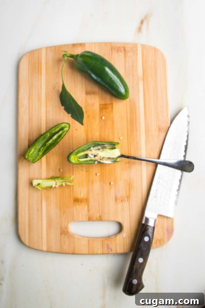 Zesty Roasted Tomatillo Salsa 5 Small cutting board with one whole jalapeno and one sliced in half lengthwise, showing a grapefruit spoon scooping out the membranes and seeds.