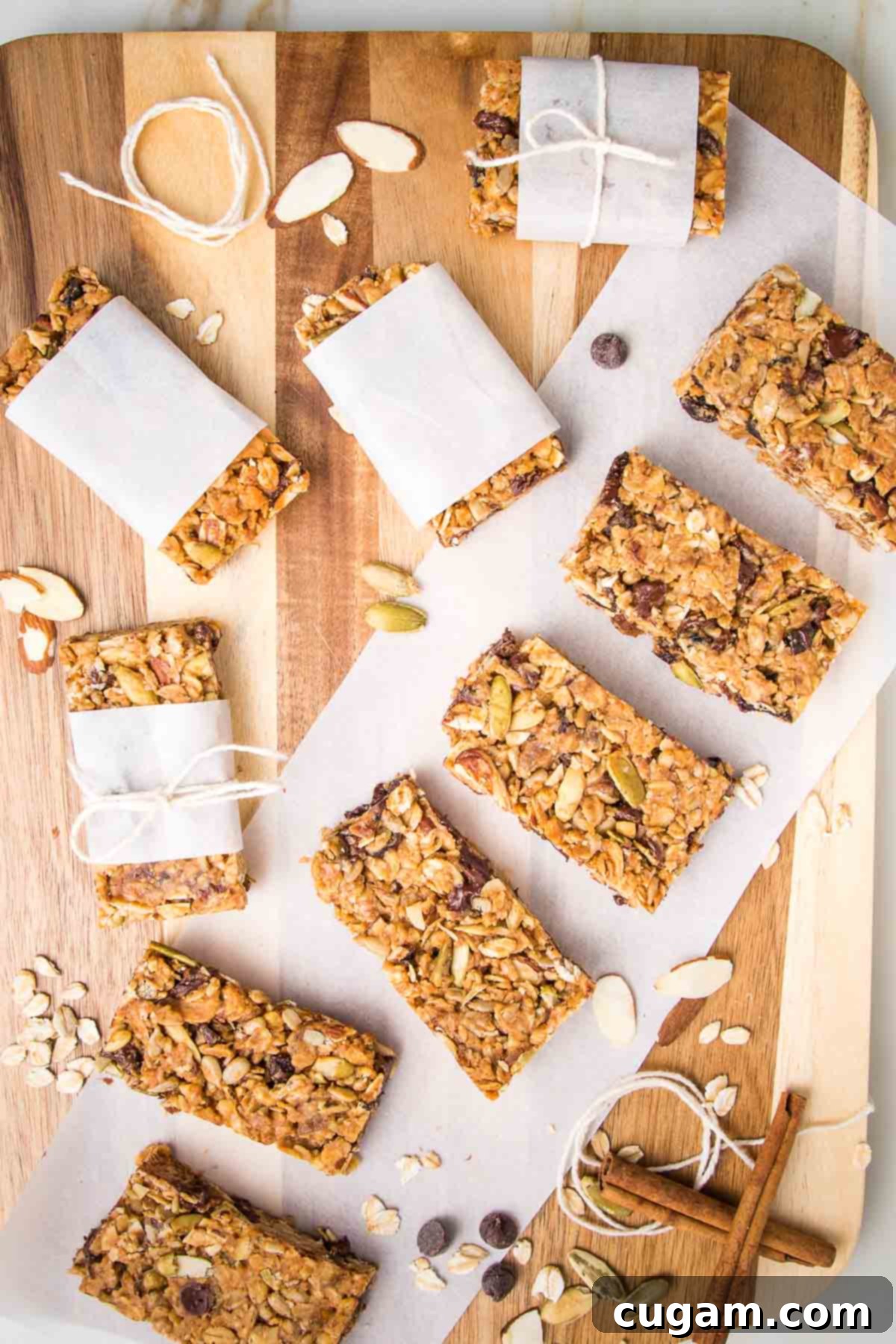 Wooden board with granola bars lined up on a piece of parchment. Also are some bars wrapped in parchment and tied with a kitchen twine. Some cinnamon stick and loose nuts and chocolate chips are strewn about.