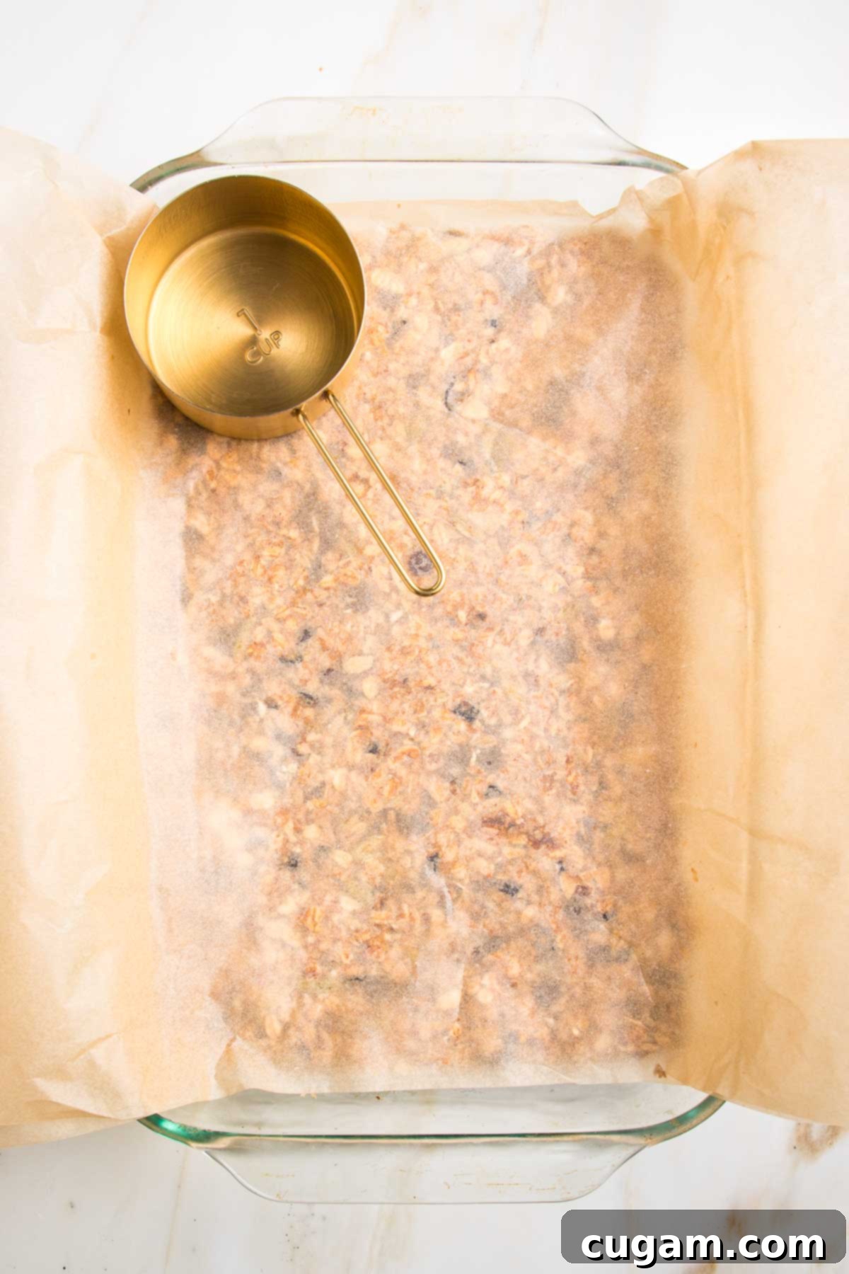 glass baking dish lined with parchment, then filled with granola bar mixture, covered with additional parchment and a gold measuring cup is patting it all down.