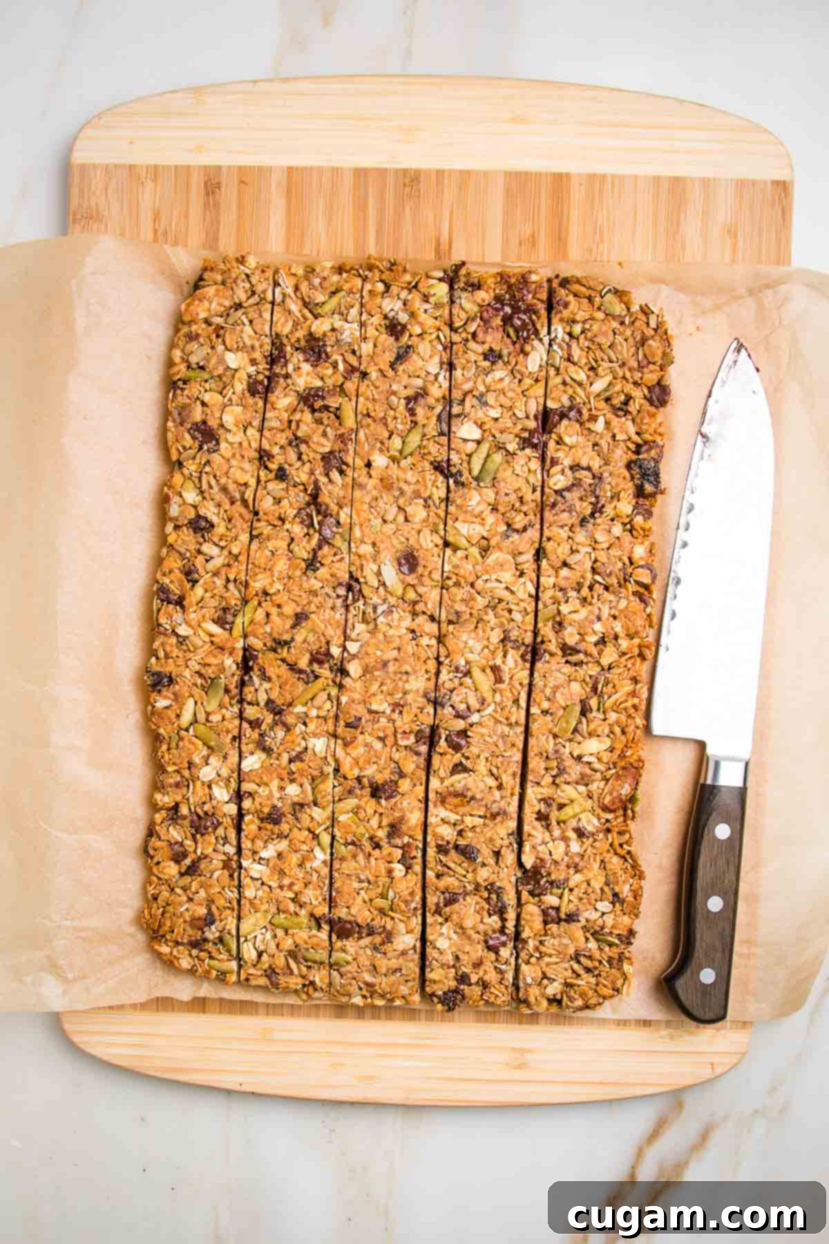 Granola bars on a piece of parchment atop a cutting board. Long thin strips have been cut with a sharp knife resting on the side.