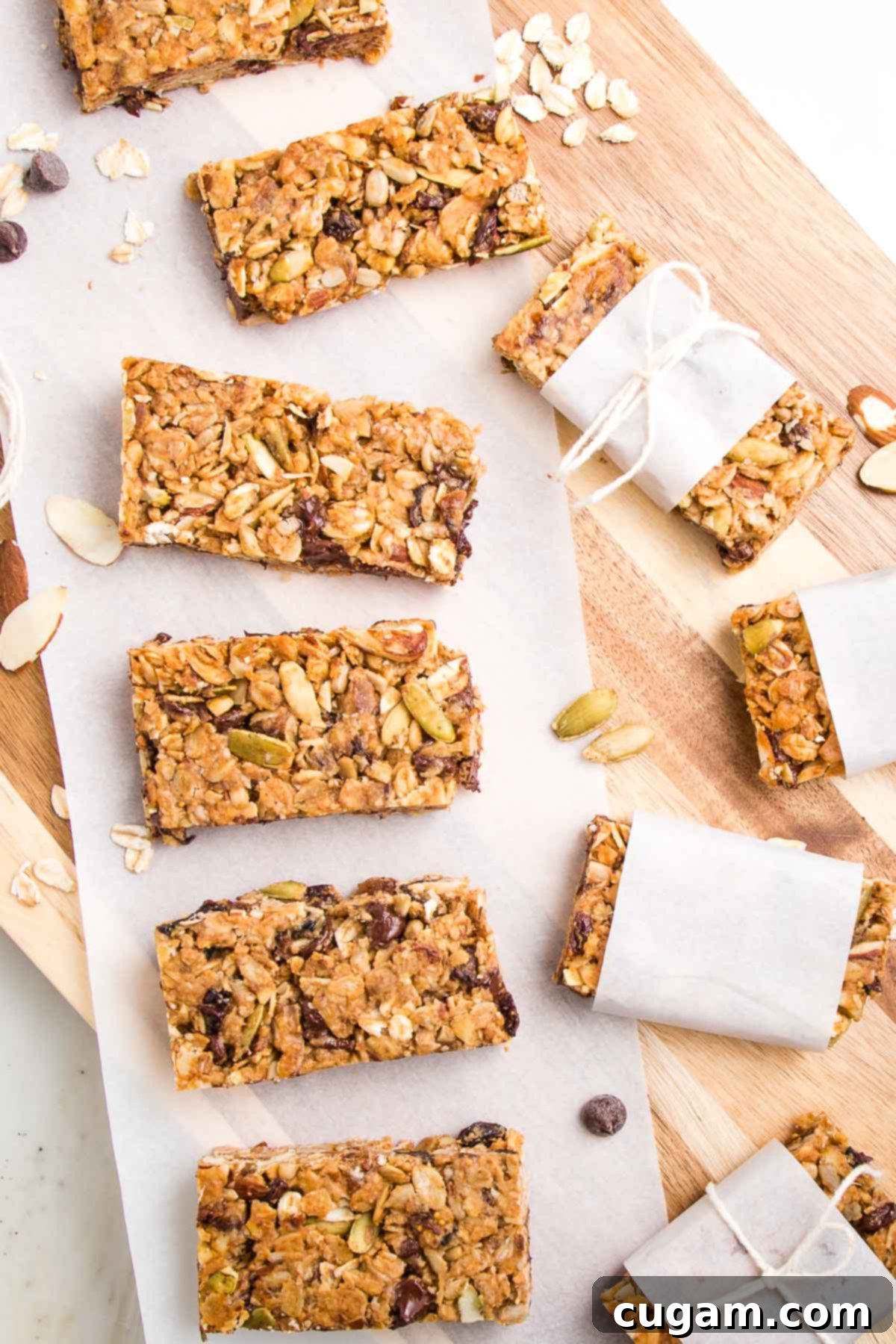 Crunchy granola bars lined up on a sheet of parchment on top of a wooden board. THere are a few wrapped in parchment strips with a kitchen twine tied around it.