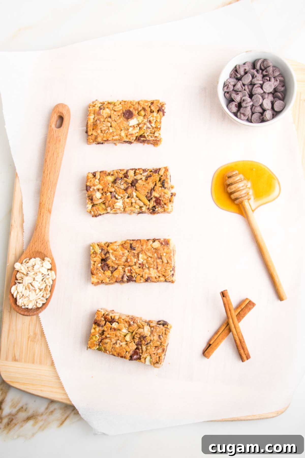 4 crunchy granola bars lined up on a piece of parchments. There is a spoon full of oats, a puddle of honey, a dish of chocolate chips and two cinnamon sticks.