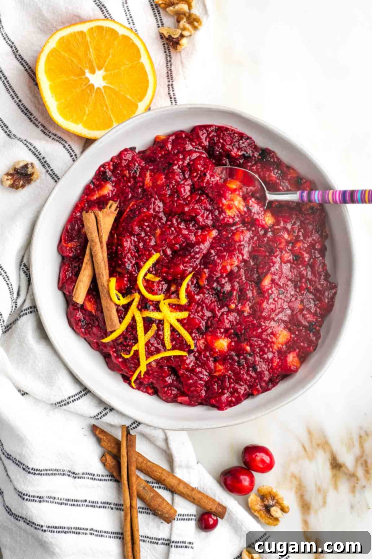Bowl of chunky cranberry sauce, surrounded by an orange, cinnamon sticks, fresh cranberries and walnuts.