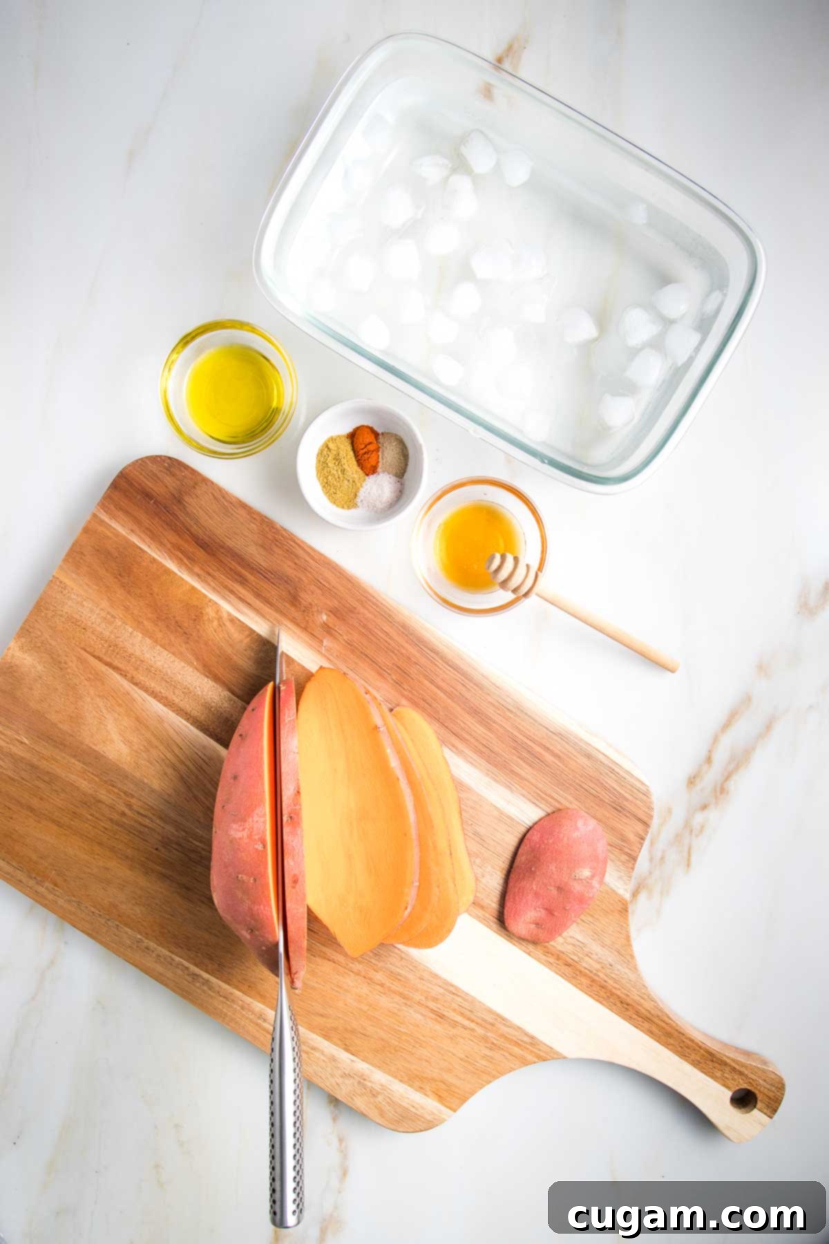 Sweet potato being sliced into ¼" long ovals. Ingredients for sweet potato fries: oil, honey, spices and a large glass dish with ice water shown.