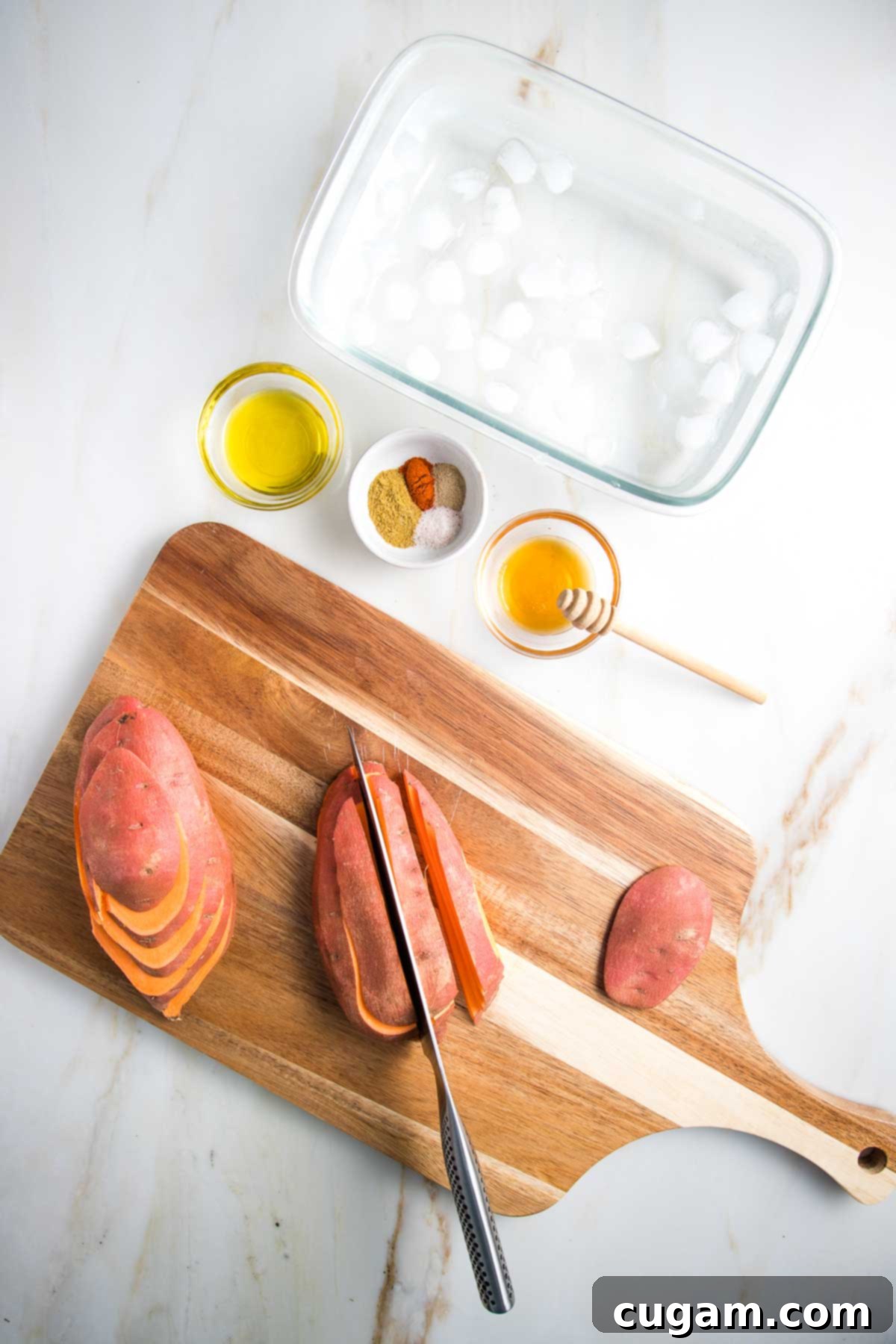 Cutting sweet potatoes into fries on a cutting board. First sliced one way, then the other. Along with glass bowls with olive oil, spices and honey, plus an ice water bath waiting to soak the fries.