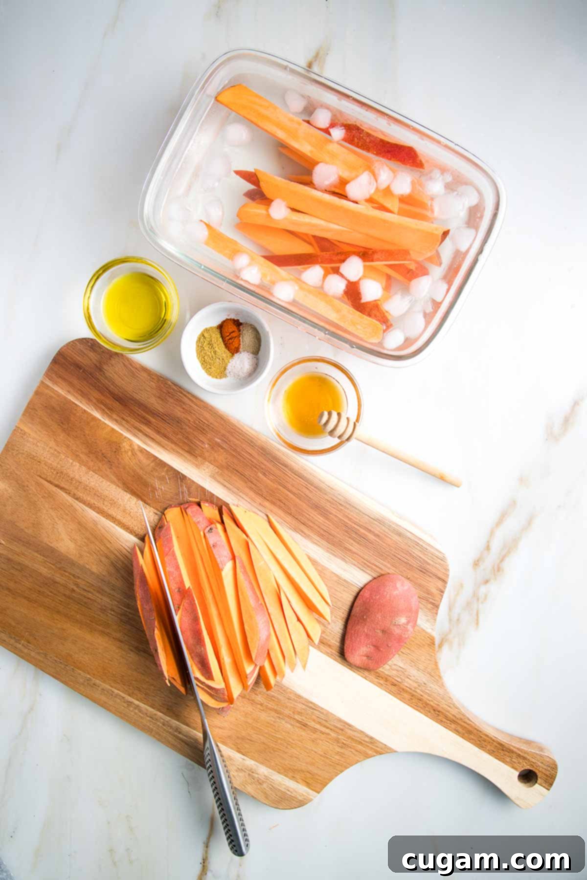 Sweet potato cut into fries on a cutting board. Some are in a large dish of ice water. Olive oil, spices and honey in small glass dishes.