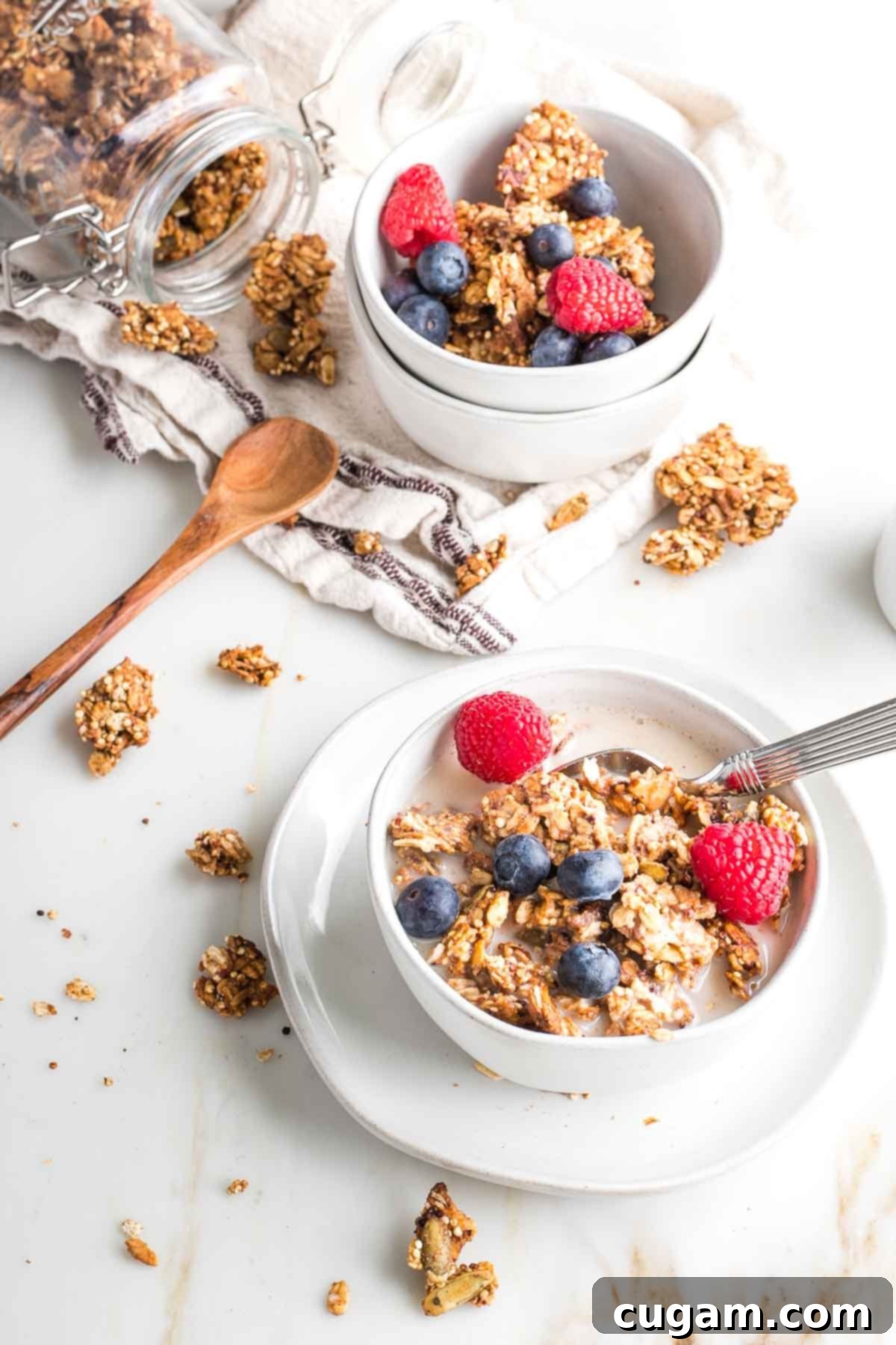 Two bowls of granola with milk and fresh berries. Clusters of granola, plus a filled jar surround the bowls.