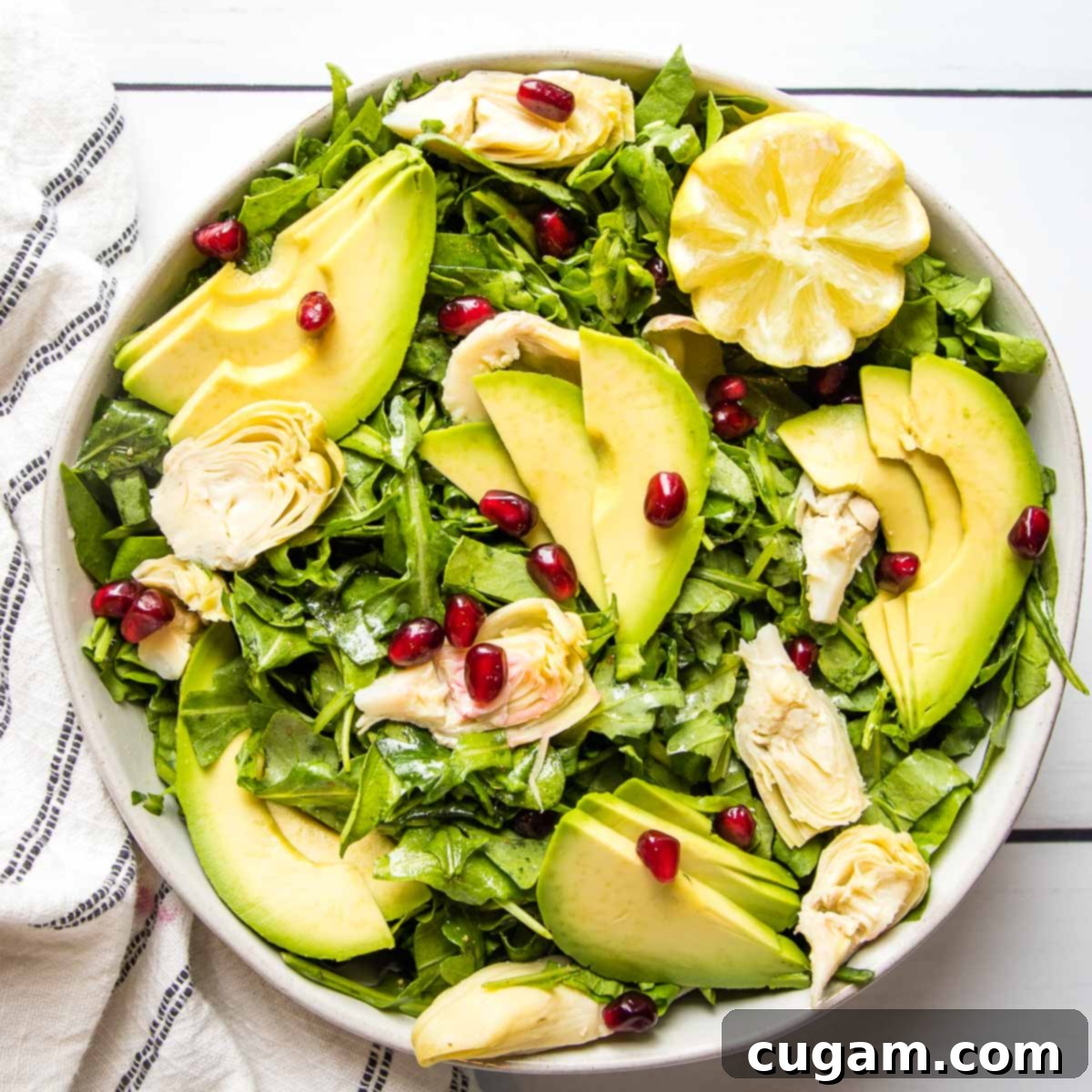 A vibrant arugula salad, elegantly presented with avocado slices, tender artichoke hearts, a sprinkle of bright pomegranate seeds, and fresh lemon wedges for garnish.