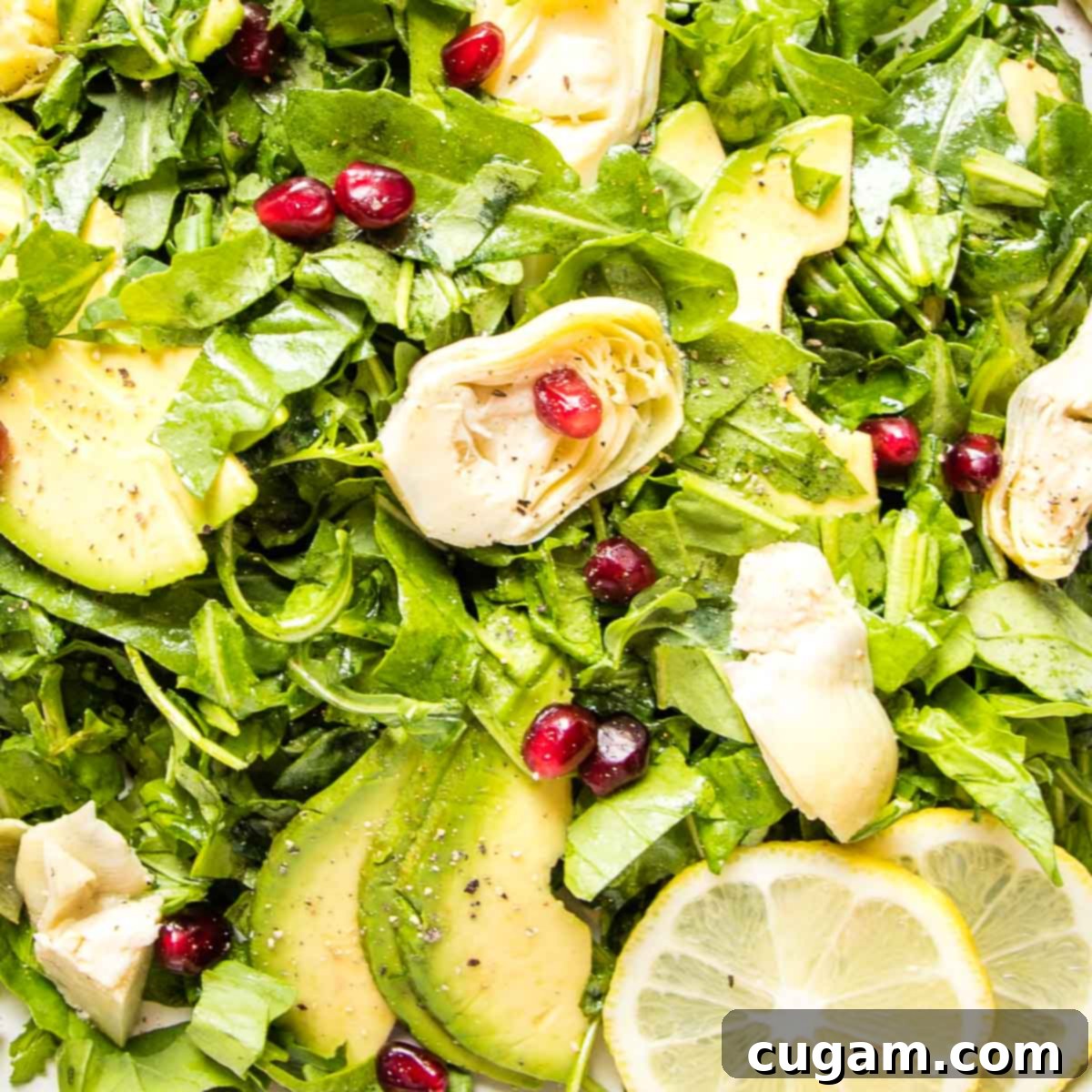 Close up view of the vibrant arugula salad, highlighting artichoke hearts, creamy avocado slices, zesty lemon wedges, and ruby red pomegranate seeds.