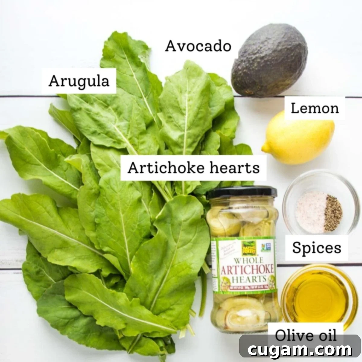 A flat lay of labeled ingredients for the arugula salad: Fresh Arugula, ripe Avocado, bright Lemon, jarred Artichoke hearts, a bowl of Spices, and a bottle of Olive oil.
