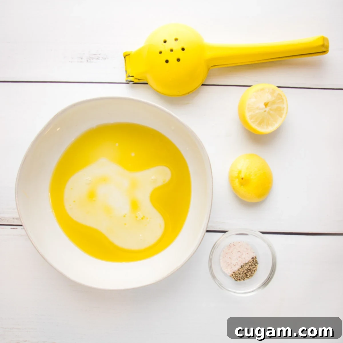 White bowl with olive oil and lemon juice that haven't been mixed, alongside two squeezed lemon halves, a yellow citrus juicer, and a small dish of salt and pepper.