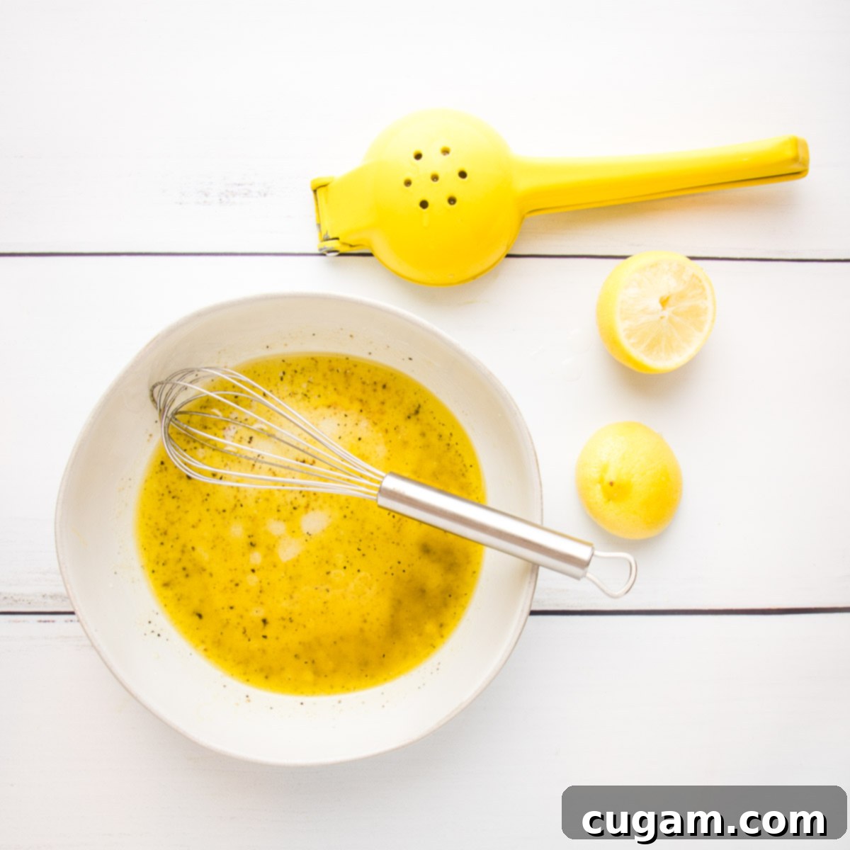 Lemon vinaigrette being emulsified in a white bowl with a whisk, with two squeezed lemon halves on the side.