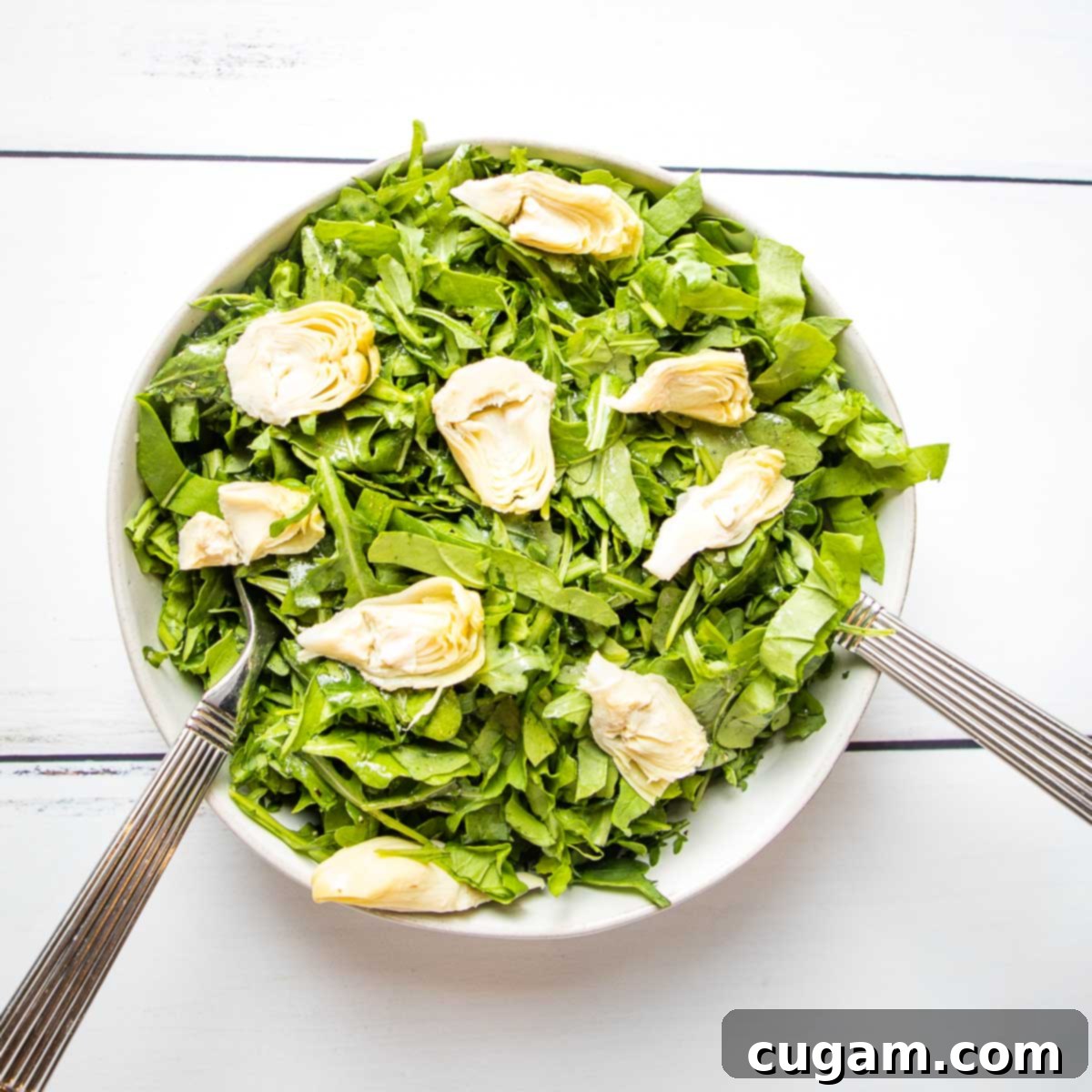 A large white bowl filled with dressed arugula, adorned with quartered artichoke hearts and silver salad tossers.