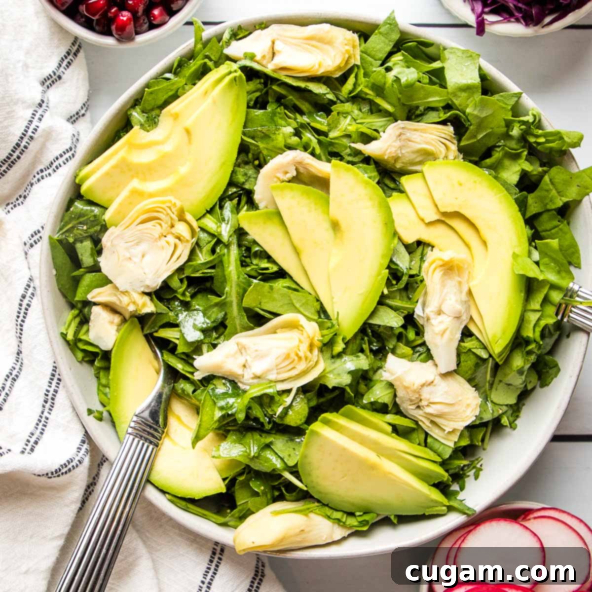 Large white bowl filled with dressed arugula, quartered artichoke hearts, and elegantly sliced avocado. Small bowls of pomegranate seeds, sliced radish, and chopped purple cabbage are arranged around the main salad bowl.
