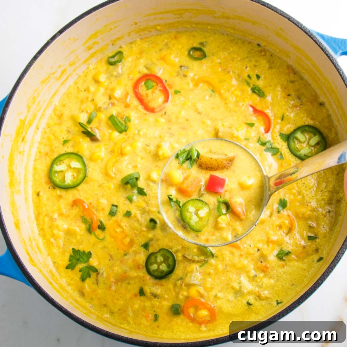 Dutch oven, filled with corn chowder, topped with colorful slices of mini bell peppers, jalapenos and fresh herbs.  The focus is on a ladle of chowder. 