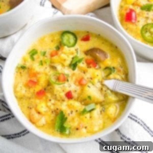 white bowl, filled with corn chowder, topped with colorful chunks of vegetables. There is a spoon I the bowl and the edges of other bowls of soup can be seen. The chowder is on top of a white linen napkin with some dark blue thin stripes.