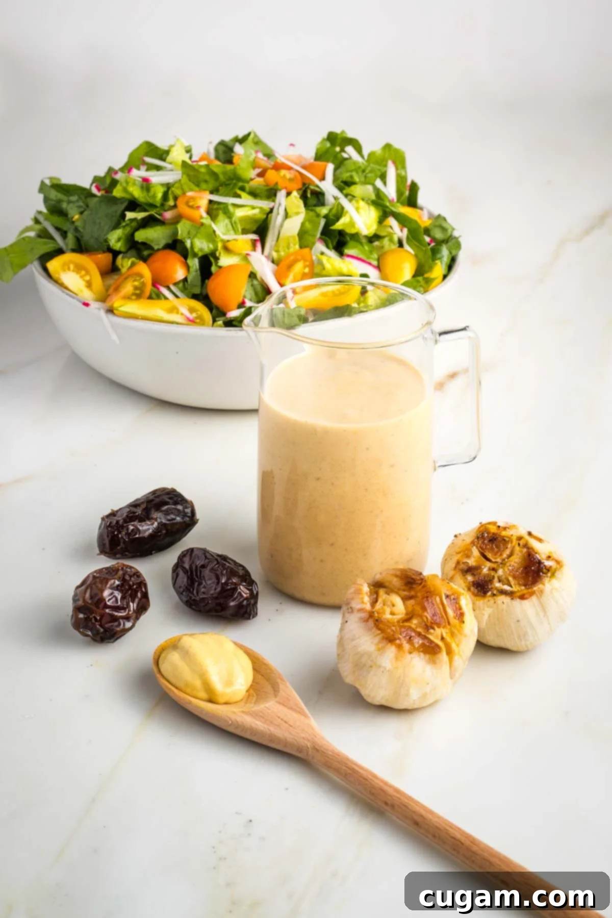Small pitcher of creamy dressing with dates, mustard and roasted heads of garlic in front of a bowl of salad.