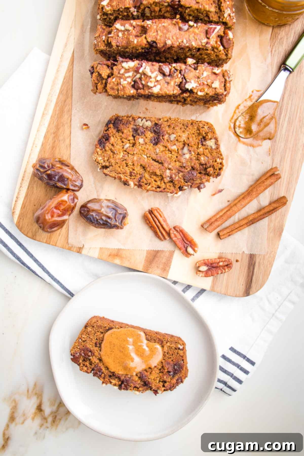 Slices of pumpkin bread with chocolate chips and pecans on a piece of parchment on a cutting board. In the foreground there is a white rimmed plate with a slice of pumpkin bread and a dollop of almond butter on top. 